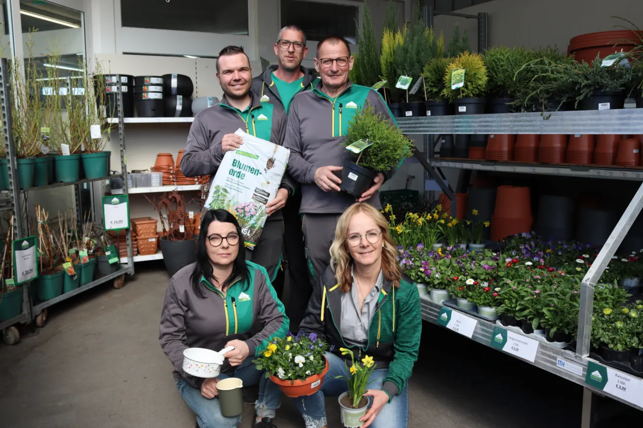 Fünf Personen posieren für ein Bild in einem Gartengeschäft. Zwei Männer stehen hinter zwei knienden Frauen. Alle tragen grüne Jacken und halten Pflanzen. Im Hintergrund befinden sich Regale mit verschiedenen Pflanzen und Töpfen.