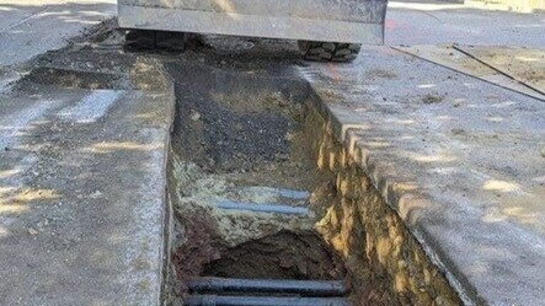 Ein Bagger steht am Straßenrand, neben einer großen ausgehobenen Öffnung im Beton. Mehrere Rohre sind in der Öffnung freigelegt.