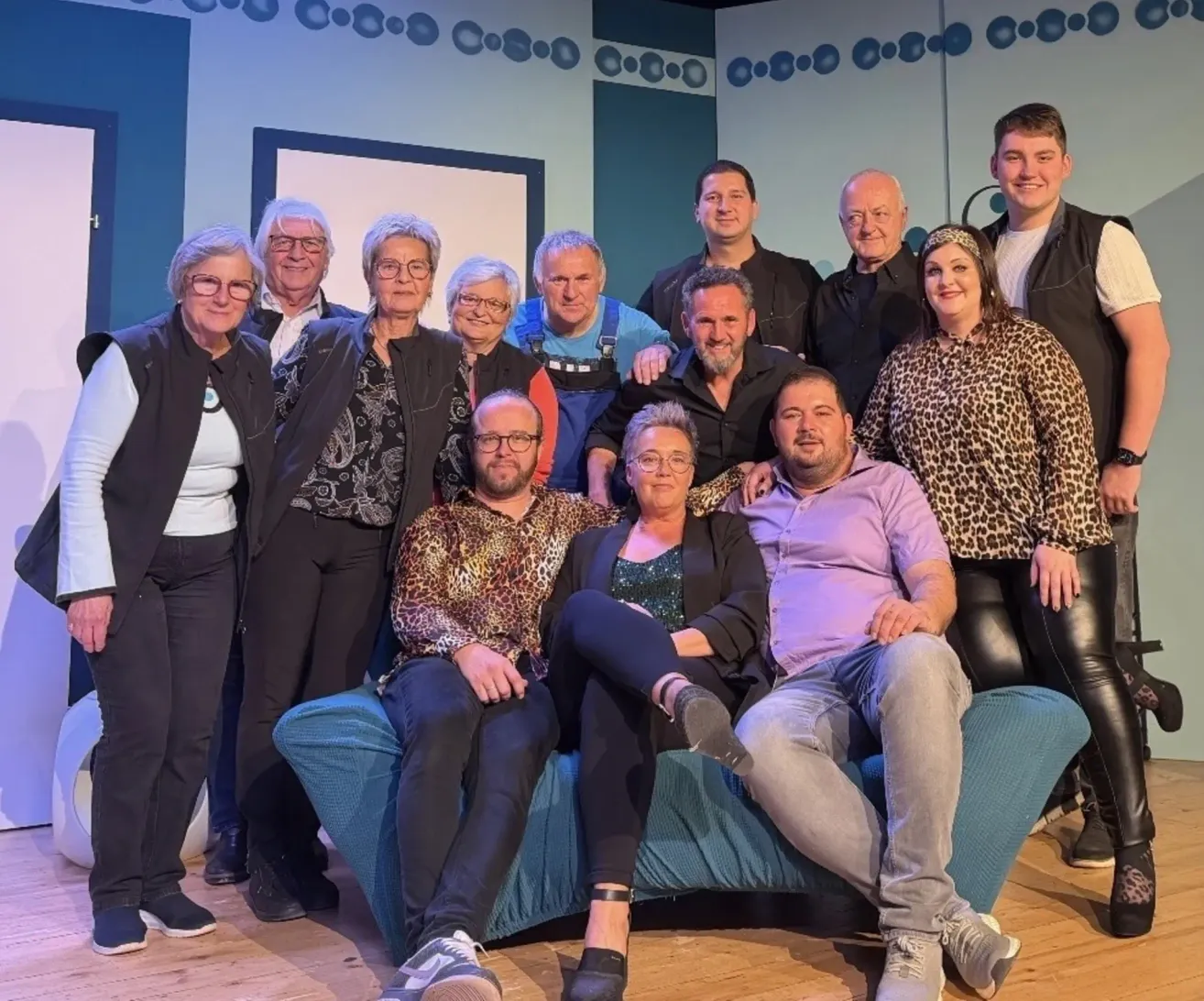 Eine Gruppe von Menschen posiert für ein Foto, einige sitzen auf einer blauen Couch, andere stehen dahinter auf einem Holzboden.