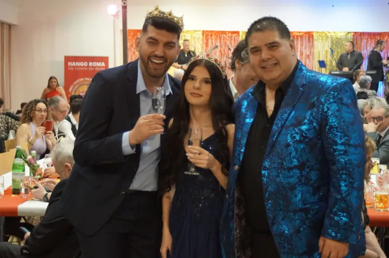 Drei Menschen in einem Party-Setting, zwei Männer und eine Frau. Der Mann auf der linken Seite trägt eine Krone und hält ein Weinglas, lächelt. Die Frau in der Mitte trägt eine Tiara und hält ein Weinglas, lächelt. Der Mann auf der rechten Seite trägt einen blauen Anzug und lächelt.