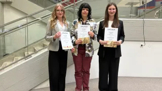 Drei Frauen stehen auf einem Betonboden, jede hält ein Zertifikat in der Hand. Sie tragen formelle Kleidung und lächeln. Hinter ihnen befindet sich eine Treppe mit einer Glasbrüstung.