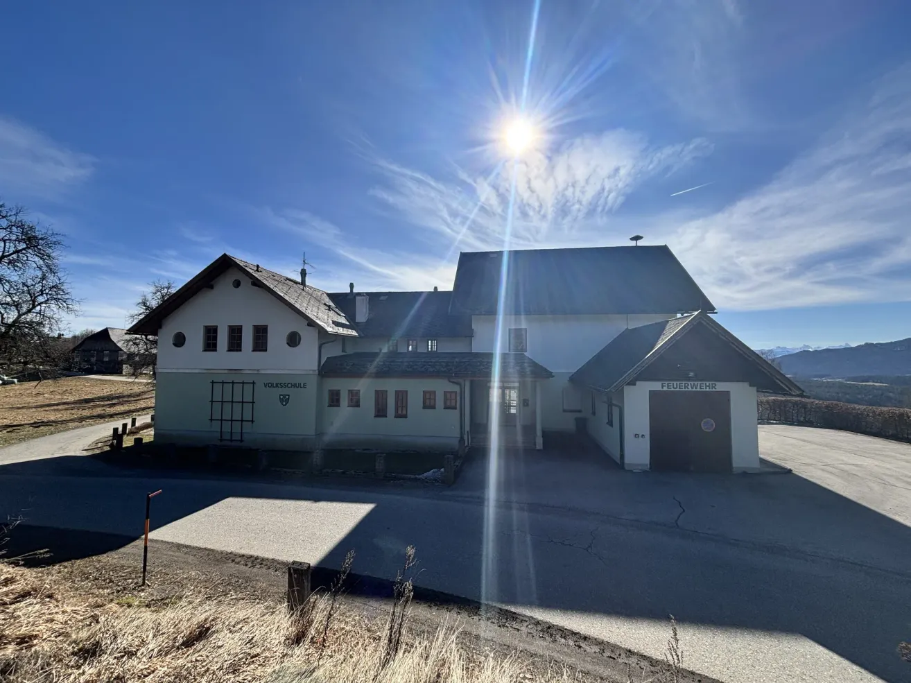 A large white building, the Volksschule, stands prominently under a bright, sunny sky. The building has multiple windows and a large garage entrance.