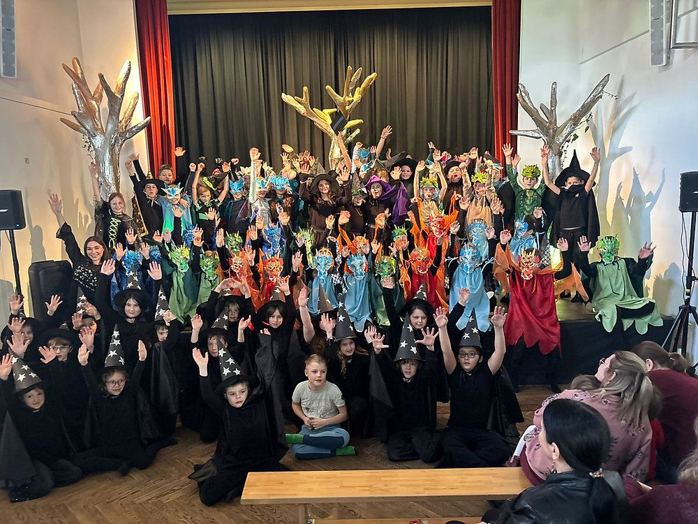 A group of children dressed in witch and wizard costumes, with hats and masks, wave and pose for a photo on a stage with trees in the background.