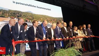 Several people in formal attire stand in a line holding a ribbon, possibly cutting it at an event. Behind them, a screen displays a landscape.