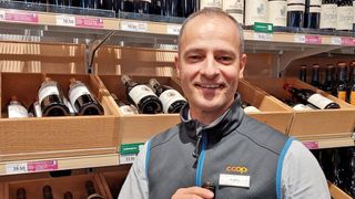 A man in a gray vest is smiling in a wine shop, holding a wine bottle. Shelves filled with wine bottles are behind him.