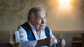 An elderly man with a mustache smiles and gestures while sitting at a table with a glass of wine.