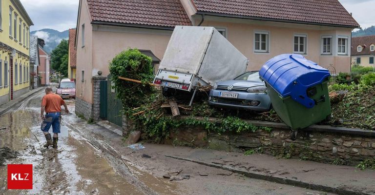 An overturned truck and a car lie on a street, both leaning against a stone wall covered in greenery. Behind them, a house with a brown roof and white windows stands.
