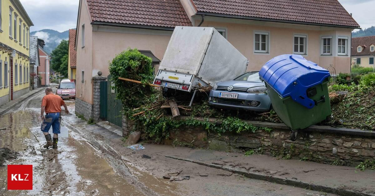 An overturned truck and a car lie on a street, both leaning against a stone wall covered in greenery. Behind them, a house with a brown roof and white windows stands.