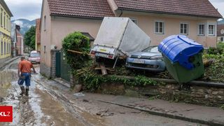 An overturned truck and a car lie on a street, both leaning against a stone wall covered in greenery. Behind them, a house with a brown roof and white windows stands.