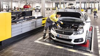 A mechanic in a yellow and gray uniform is working on a white Peugeot car in a well-lit garage. Other cars are parked or being serviced.