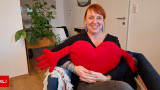 A woman with red hair and tattoos is sitting on a couch, holding a large red heart-shaped pillow with gloves. She is smiling and seems to be posing for a photo.