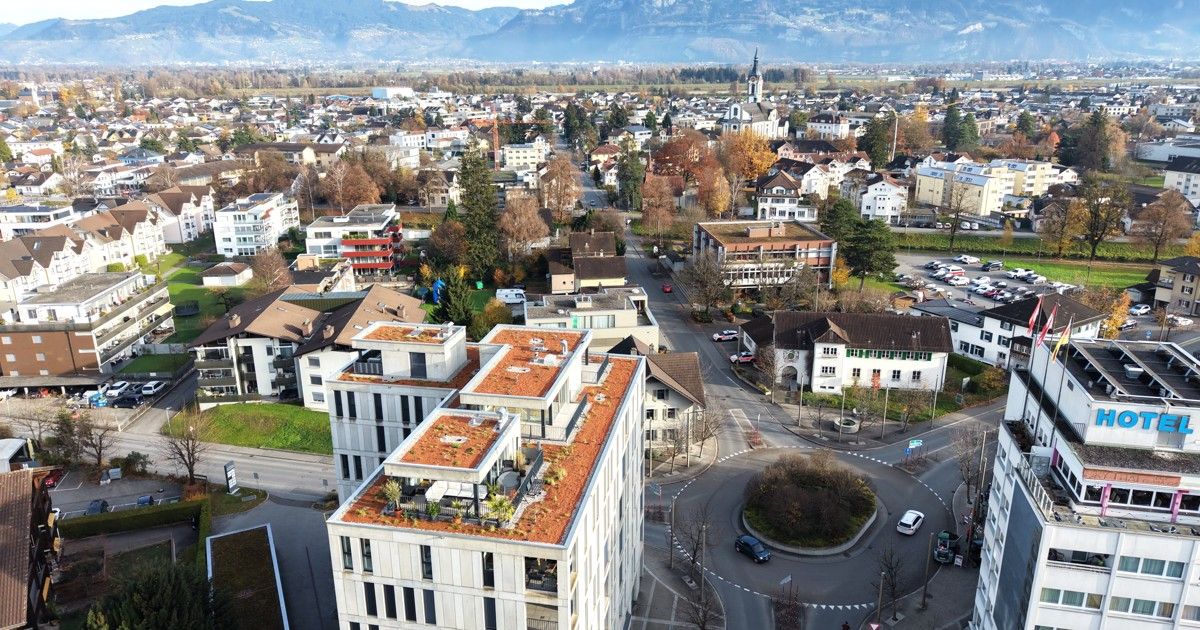 An aerial view of a modern urban area with various buildings and a roundabout. The area is surrounded by lush greenery and trees. In the distance, there are mountains.