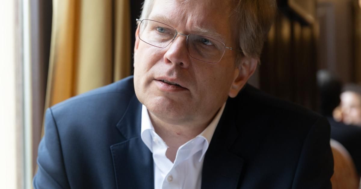 A man in a navy blue suit and white shirt sits inside, looking thoughtfully to his right.