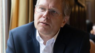 A man in a navy blue suit and white shirt sits inside, looking thoughtfully to his right.