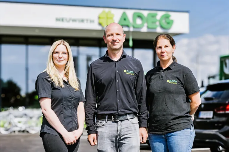 Three people in black shirts, standing in front of a building with the sign ADEG. The man in the middle wears a belt. The women on the left and right have glasses and short hair, respectively.