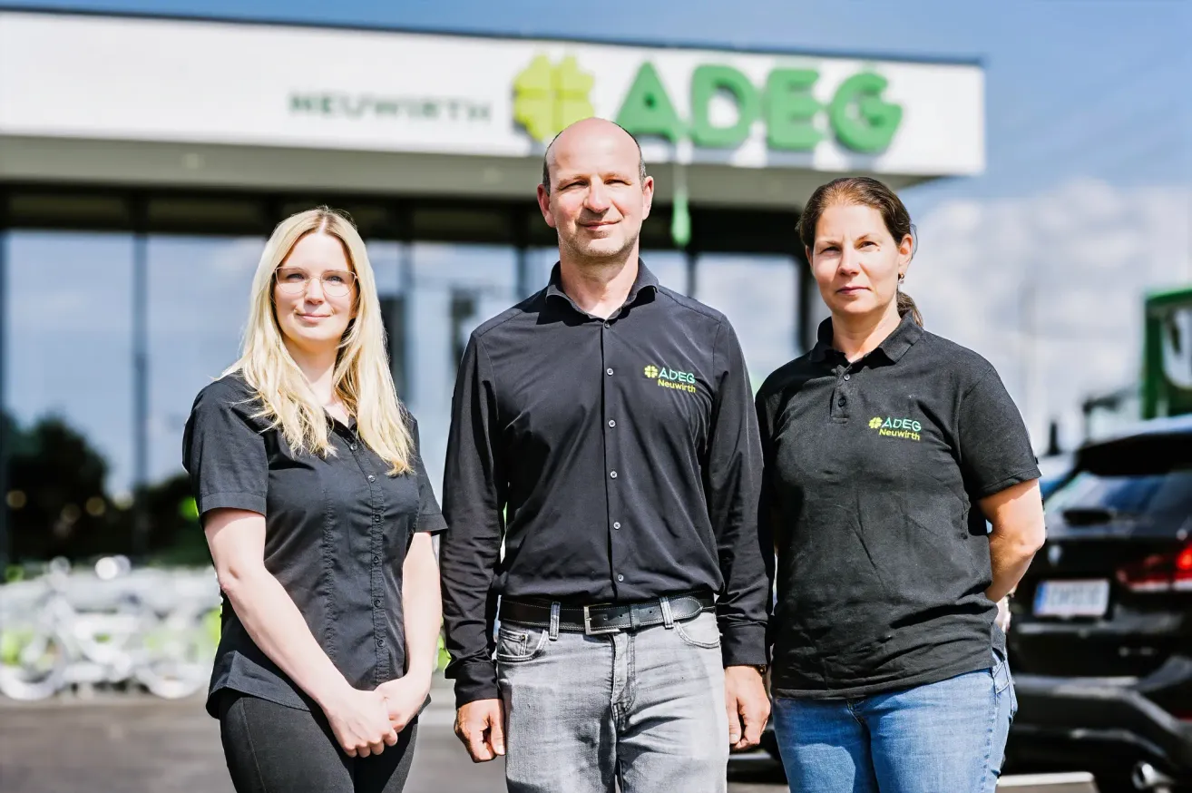 Three people in black shirts, standing in front of a building with the sign ADEG. The man in the middle wears a belt. The women on the left and right have glasses and short hair, respectively.