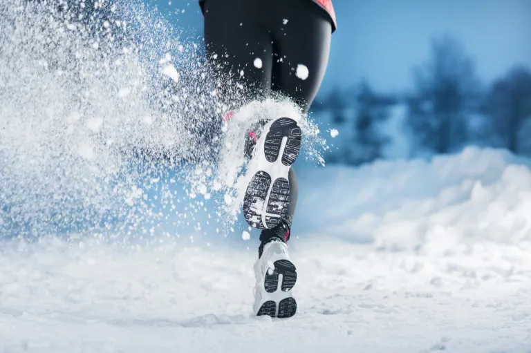 A person is running in the snow, causing a splash of snow around their feet. They are wearing black leggings and sneakers.