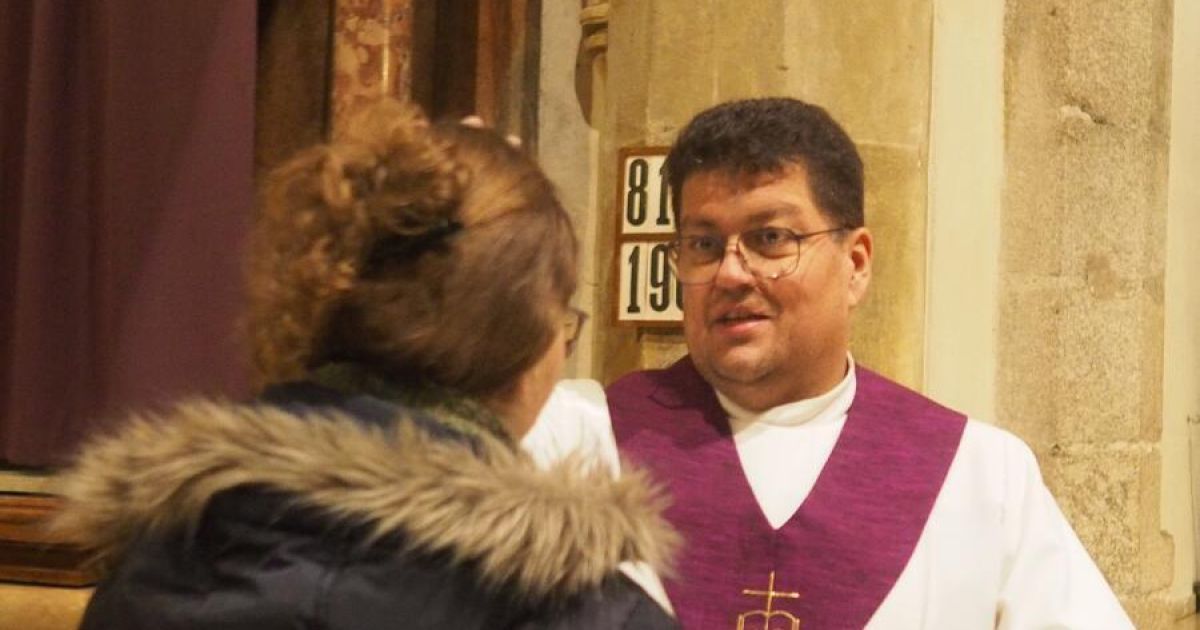 A priest in a purple vestment stands behind a woman with glasses and a fur-lined jacket. They are near a building with a number 19 sign.