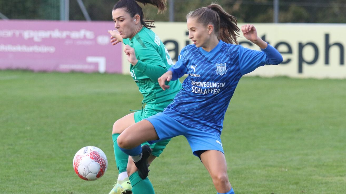Two women soccer players are on a field. One is in a green jersey and the other in a blue jersey with the word Schlaffer on it. They are both trying to kick a soccer ball.