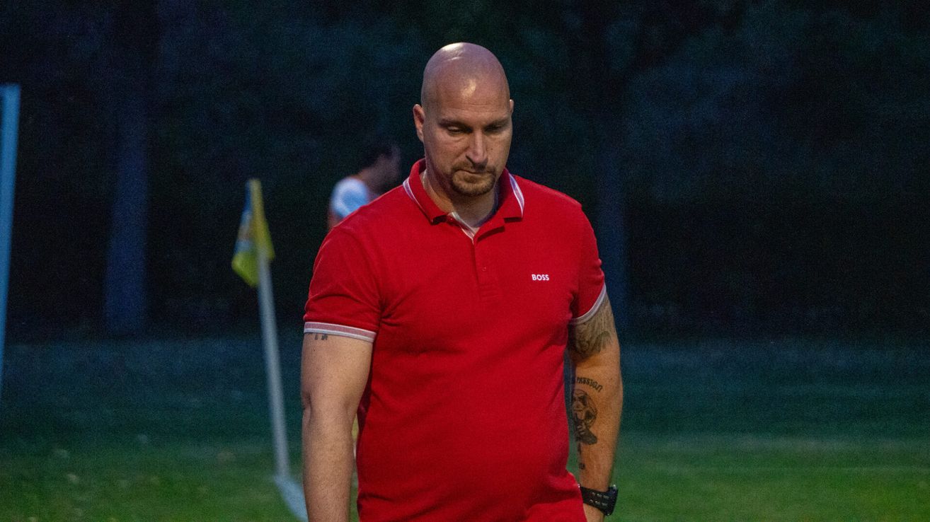 A bald man wearing a red polo shirt with the word 'BOSS' on it, walks on a grassy field under a dark sky. He has tattoos on his arms.
