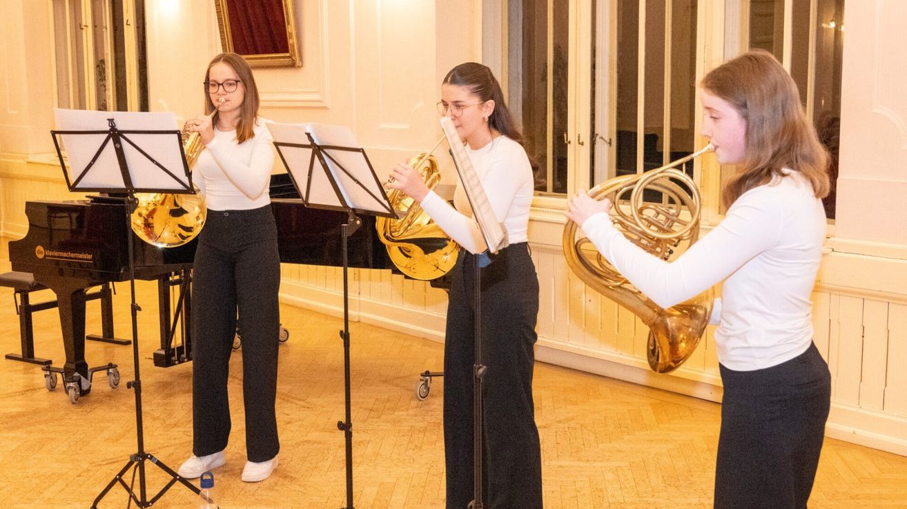Three women in matching white tops and black pants play brass instruments in a room with a piano and wooden floor.