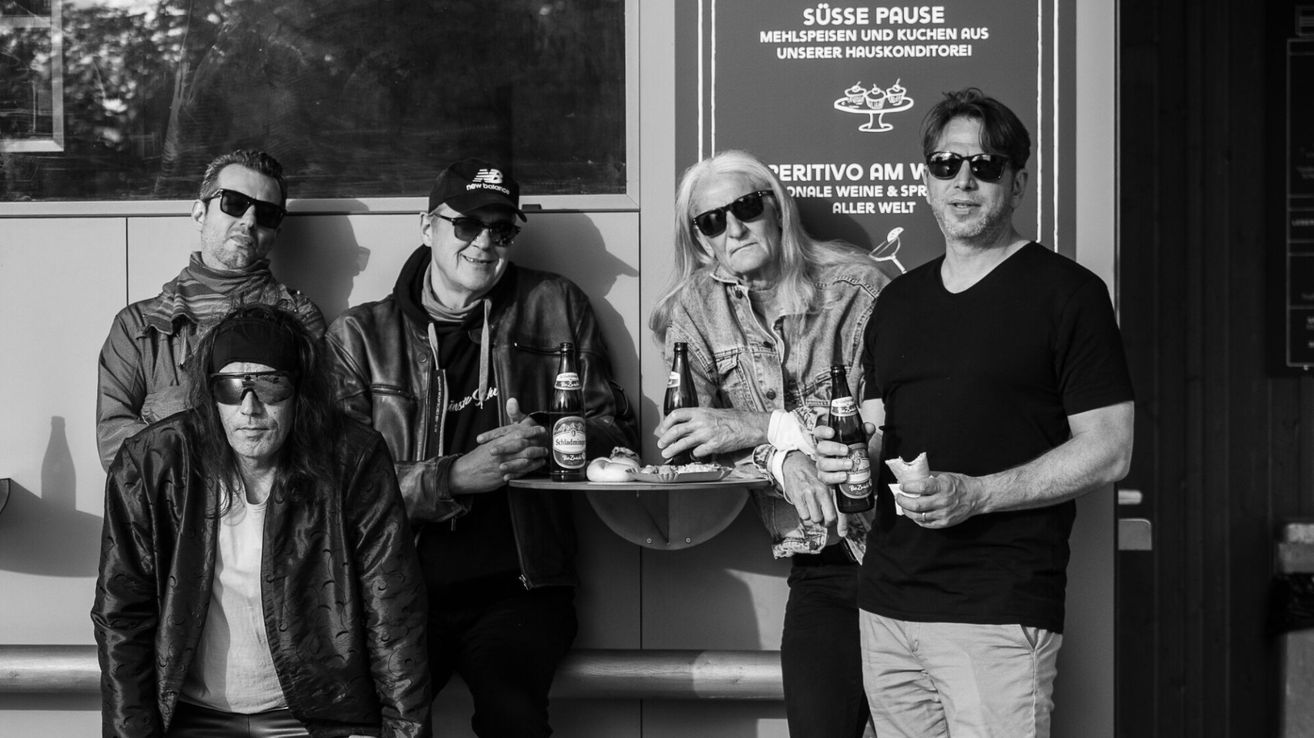 Four men stand together, smiling, in front of a wall. They hold bottles and food. A sign in the background reads 'Süsse Pause'.