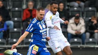 Two soccer players in action during a match, one in blue and the other in white. The blue-clad player looks determined, while the white-clad player is focused. Spectators watch from the stands.