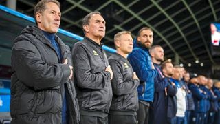 A group of men in sports attire stand together, possibly at a sports event, with focused expressions and arms crossed.
