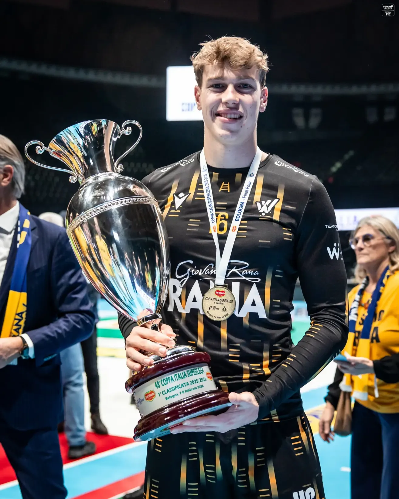 A man holds a large silver trophy in his hands, smiling. He wears a black sports uniform with the number 6 on it. A woman in a yellow shirt stands behind him.