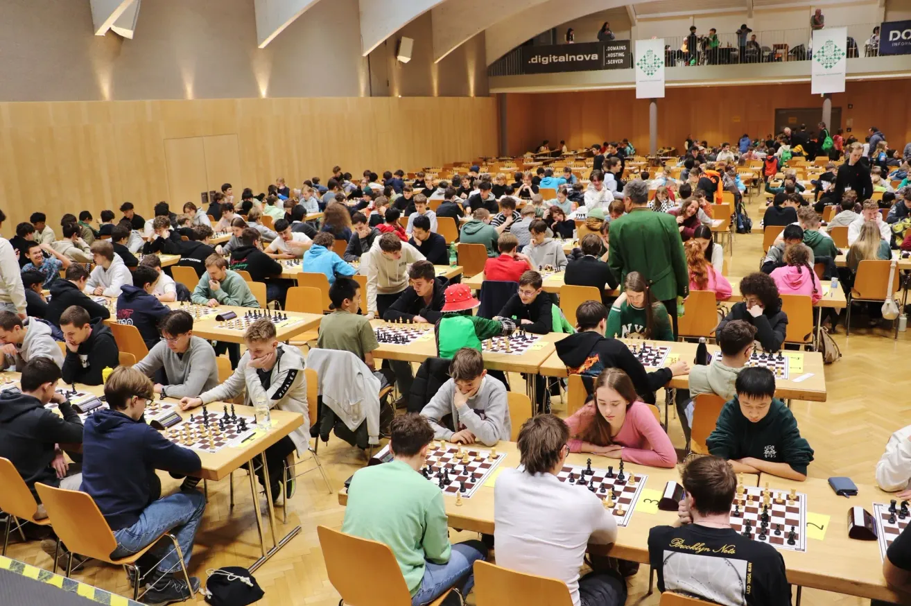 A large group of people are sitting at tables in a hall playing chess. The tables have chess boards and pieces. Some people are wearing jackets and hoodies. An adult male is standing in front of a banner.
