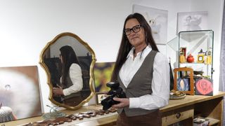 A woman with glasses stands in front of a gold mirror holding a Sony camera. She wears a white shirt and a gray vest. The reflection shows her holding a bottle.