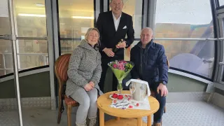 Three people pose for a photo, two sitting and one standing. A woman holds flowers and a man holds a bottle. A round table has a wine glass, ice bucket, and a plate.