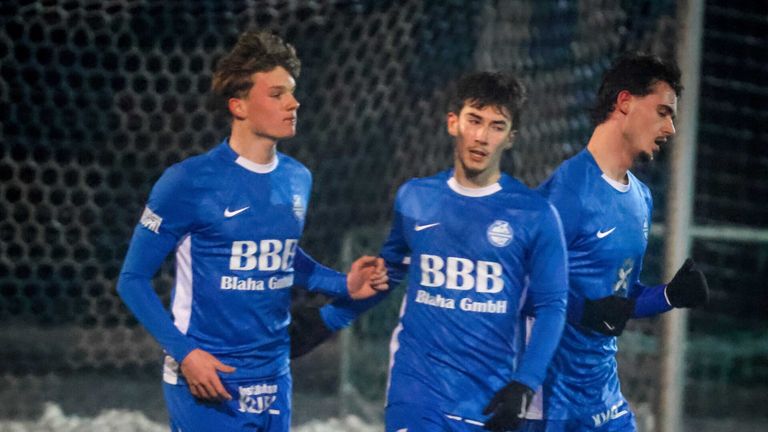 Three young soccer players in blue jerseys with white accents are standing together, likely in a stadium.