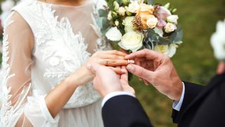 A groom places a ring on the bride's finger during a wedding ceremony, with a floral bouquet in the background.