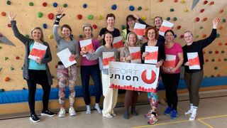 A group of women stands in front of a climbing wall, holding certificates. They are smiling and posing for a photo.