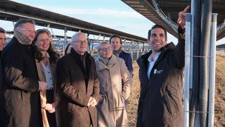 A group of people, including two men and two women, stand in a field under a solar panel. The man on the right wears a jacket with a logo.
