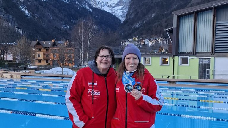Two women stand in front of a pool, both wearing red jackets with medals. They are smiling, and one holds two medals. The background shows a mountainous area and a building.