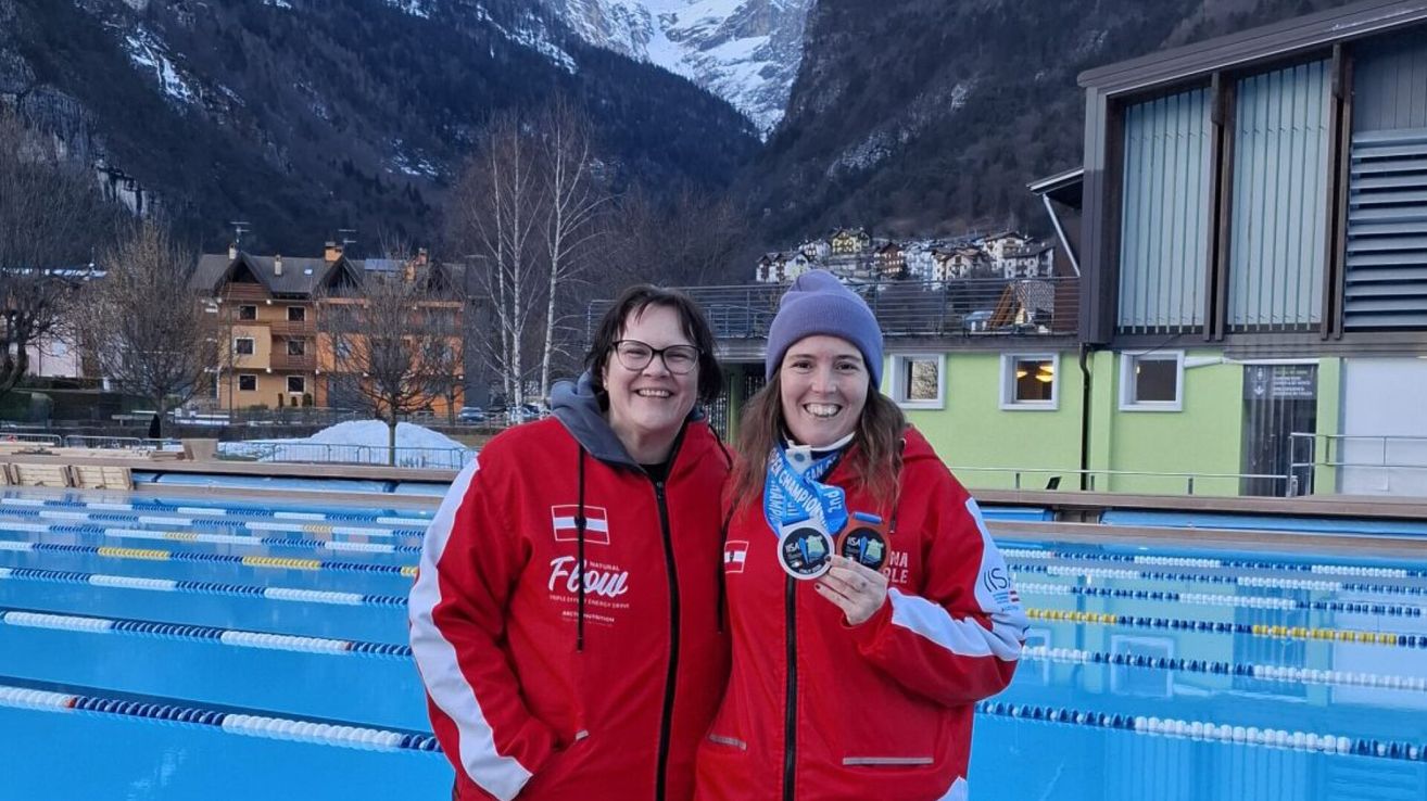 Two women stand in front of a pool, both wearing red jackets with medals. They are smiling, and one holds two medals. The background shows a mountainous area and a building.