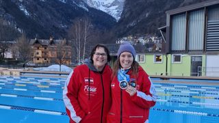 Two women stand in front of a pool, both wearing red jackets with medals. They are smiling, and one holds two medals. The background shows a mountainous area and a building.