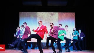 A group of men in colorful suits perform a synchronized dance on a stage with a large screen backdrop.