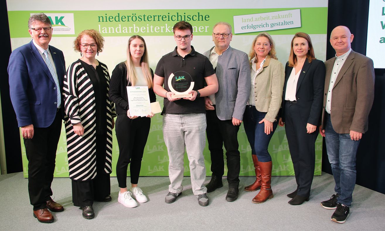 A group of people, possibly colleagues, stand in front of a green backdrop with text, posing for a photo. The man in the middle holds an award and certificate. The woman on the left wears a black and white striped coat.