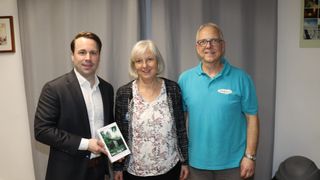 Three people stand together smiling. A man in a suit holds a book titled 'Der Tag X'. The woman next to him wears a patterned top and a necklace. The man on the right wears a blue polo shirt.