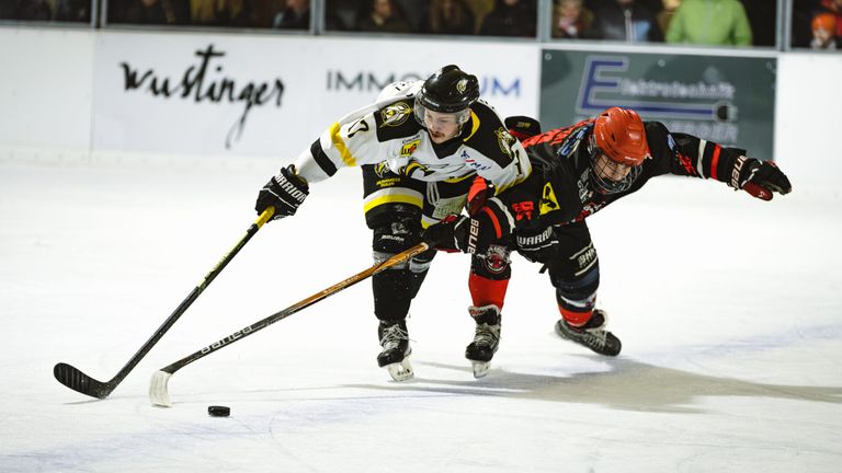 Two hockey players on an ice rink, one in white and the other in red, are in action, one skating forward while the other leans in to defend.