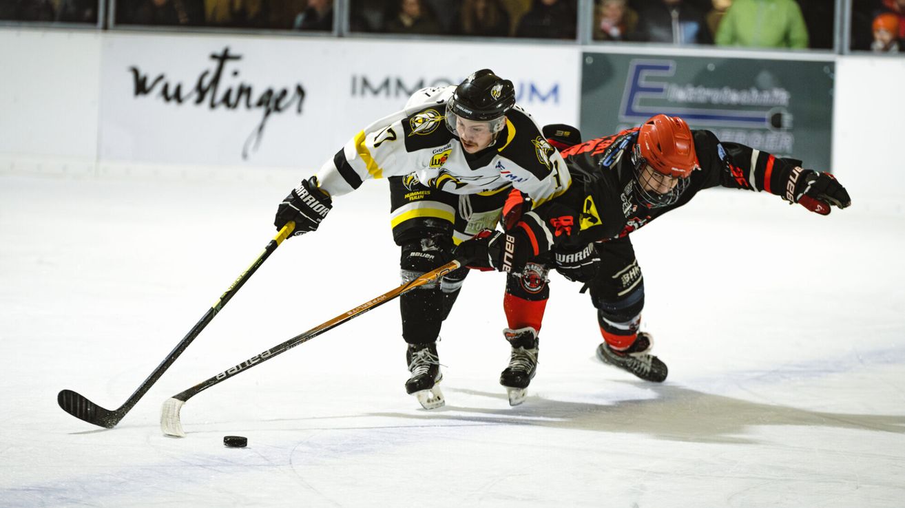 Two hockey players on an ice rink, one in white and the other in red, are in action, one skating forward while the other leans in to defend.
