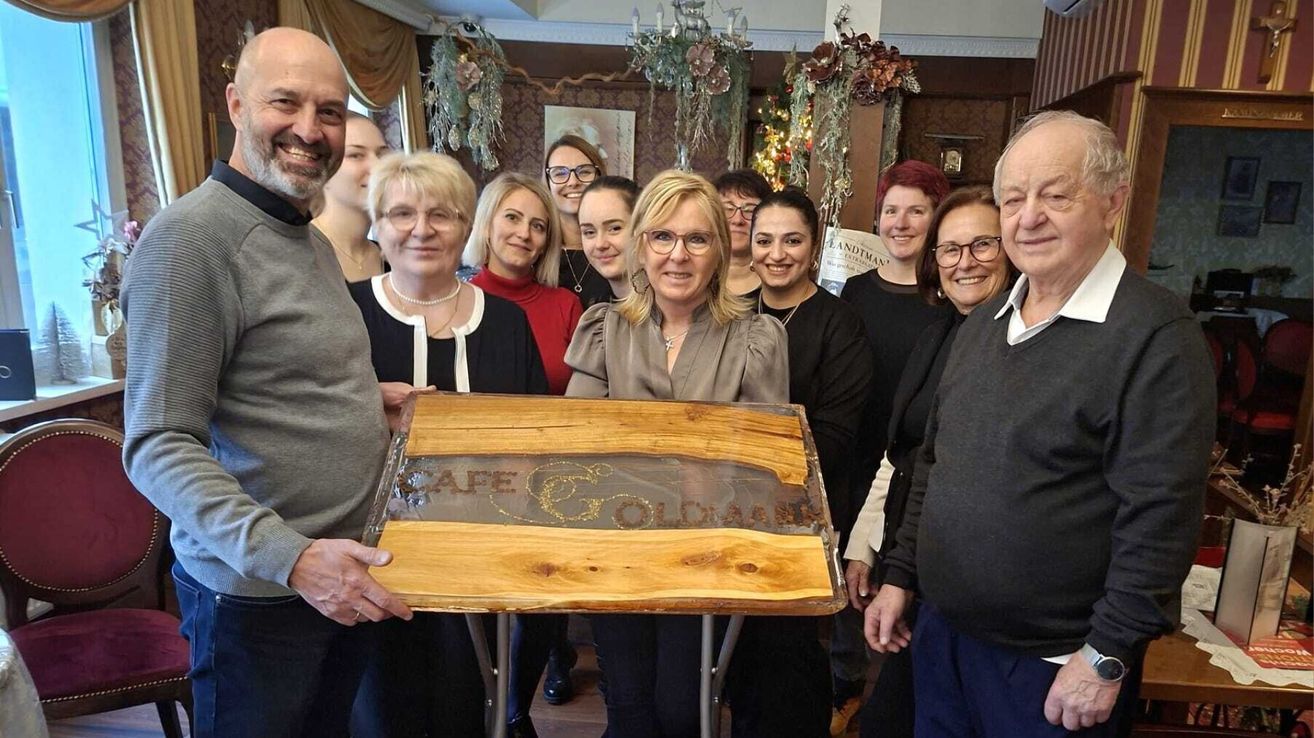 A group of people, mostly women, stand around a wooden table, holding a plaque that says 'Cape Goldmine'. They are smiling and appear to be posing for a photo. They are dressed casually. Behind them, there are decorations and a chandelier.