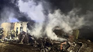 Das Bild zeigt ein ausgebranntes Gebäude, aus dem dicker Rauch aufsteigt. Die Überreste des Gebäudes stehen inmitten verkohlter Trümmer.