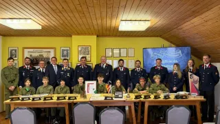 A group of people in uniforms are posing for a photo. They are standing and sitting around a table with trophies. Some of them are wearing hats. Behind them are men in uniforms standing. There are picture frames hanging on the wall. There is a large screen on the wall.