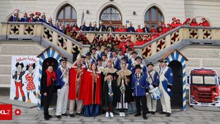 A large group of people in traditional costumes poses in front of a historic building with ornate staircases and heart-shaped decorations. Some hold musical instruments and flags.