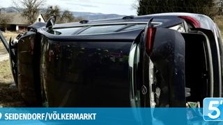 A black vehicle is overturned, with emergency personnel around it. The vehicle has a Mercedes logo on the door. In the background, there is a house and mountains.