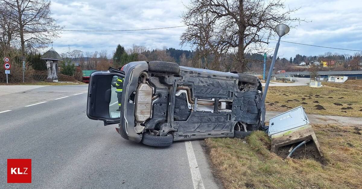 An overturned vehicle lies on its side on the roadside. The car's underside is exposed, and its tires are detached. A person stands near the car. Trees and a streetlight are visible in the background.